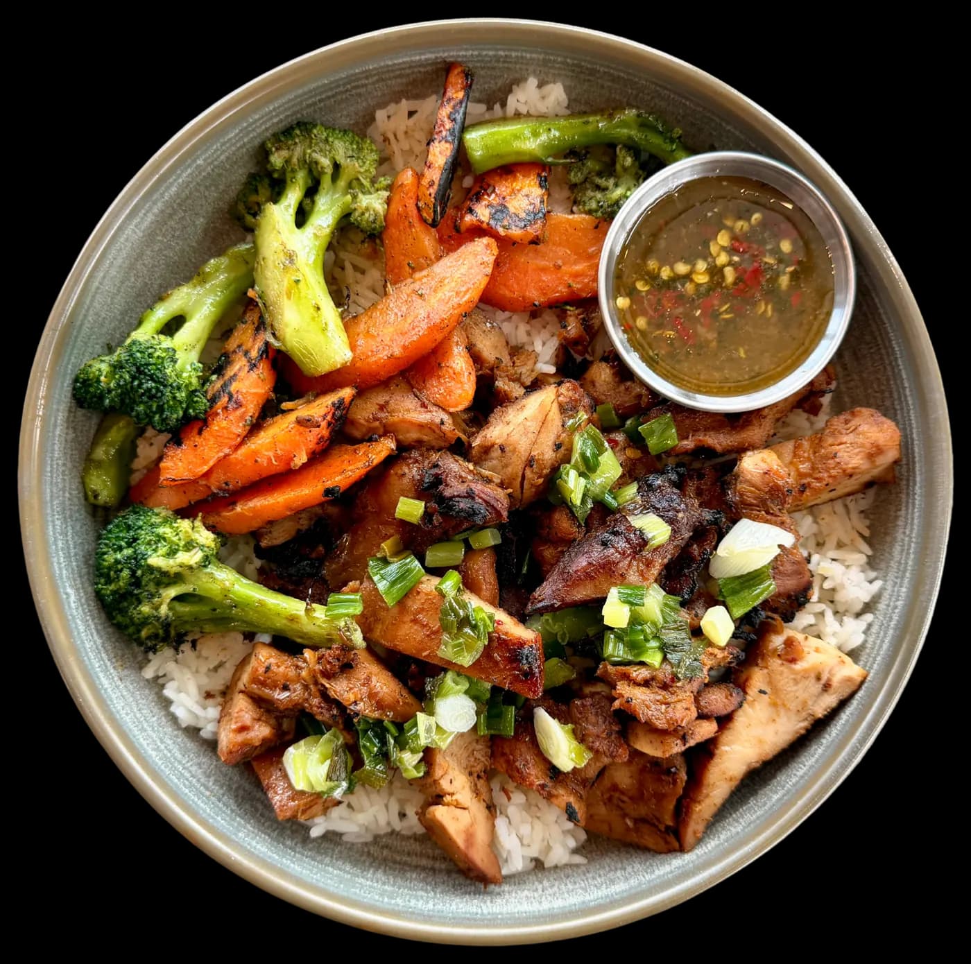 A NOTO Grilled Chicken Rice Bowl with charred broccoli, roasted carrots, spring onion and a side of dipping sauce.